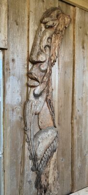 Tall carved post with a face leaning against a timber wall